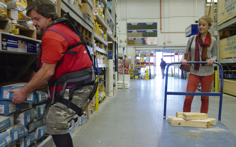 Lowe's Home Improvement associate on sales floor lifting heavy object for female customer while wearing soft robotic exoskeleton for assistance.